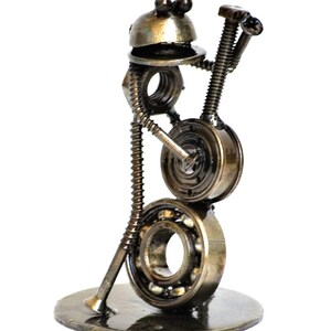 May include: A handcrafted metal sculpture of a musician playing a banjo. The sculpture is made from various metal hardware pieces, including nuts, bolts, and bearings. The banjo player is standing on a round metal base.