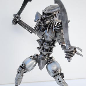 May include: A silver metal sculpture of a Predator from the movie franchise. The sculpture is made of various metal parts, including nuts, bolts, and screws. The Predator is holding two swords in its hands and is in a fighting stance.