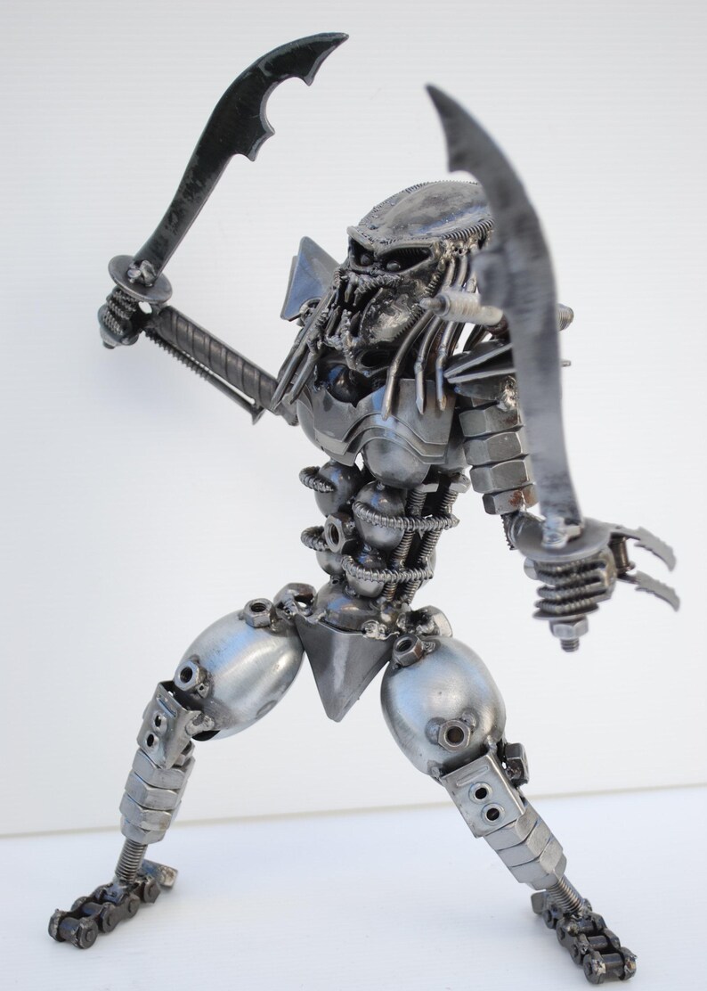 May include: A silver metal sculpture of a Predator from the movie franchise. The sculpture is made of various metal parts, including nuts, bolts, and screws. The Predator is holding two swords in its hands and is in a fighting stance.