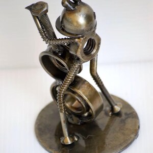 May include: A handcrafted metal sculpture of a whimsical figure, assembled from various hardware components. The sculpture features a round base, a body made of a bearing, and a head with two small protrusions. The figure appears to be playing a musical instrument.