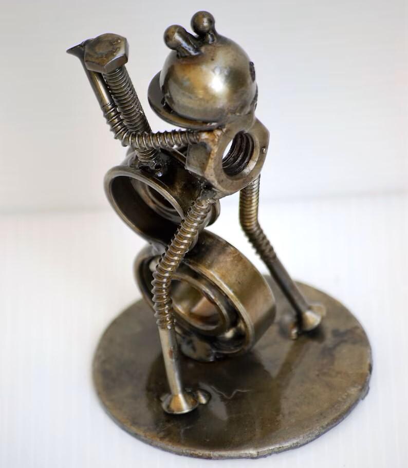 May include: A handcrafted metal sculpture of a whimsical figure, assembled from various hardware components. The sculpture features a round base, a body made of a bearing, and a head with two small protrusions. The figure appears to be playing a musical instrument.