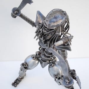 May include: A metal sculpture of a Predator, a fictional alien hunter, made from recycled metal parts. The sculpture is posed in a hunting stance, with one arm raised and a weapon in its hand. The Predator's head is detailed with a helmet and dreadlocks.
