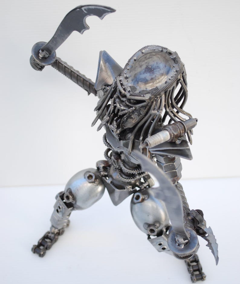 May include: A metal sculpture of a Predator, a fictional alien hunter, made from recycled metal parts. The sculpture is posed in a hunting stance, with one arm raised and a weapon in its hand. The Predator's head is detailed with a helmet and dreadlocks.