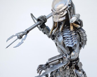 May include: A silver metal sculpture of a Predator from the movie franchise. The Predator is holding a spear in its right hand and has a detailed, articulated body.