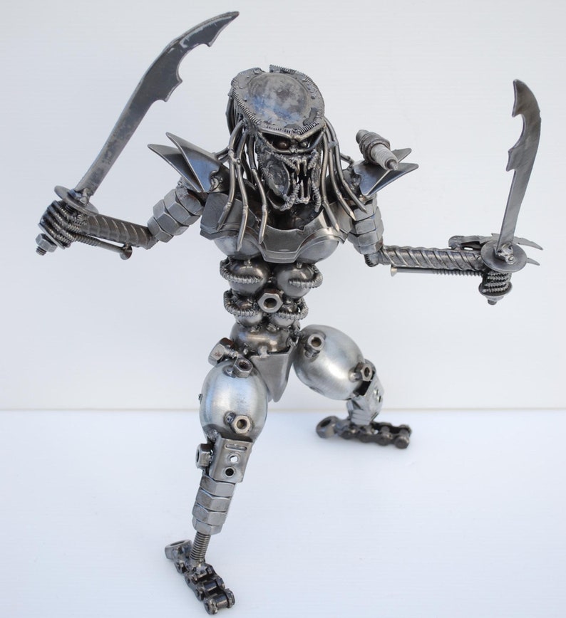 May include: A silver metal sculpture of a Predator from the movie franchise. The Predator is holding two swords in its hands and is in a fighting stance. The sculpture is made of various metal parts, including nuts, bolts, and screws.