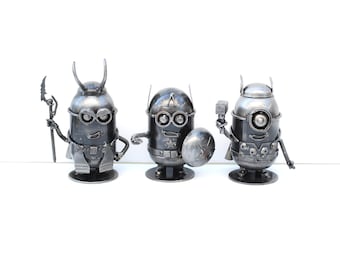 May include: Three metal figurines of Minions dressed as superheroes. The Minion on the left is dressed as Thor, the middle Minion is dressed as Captain America, and the Minion on the right is dressed as Iron Man.