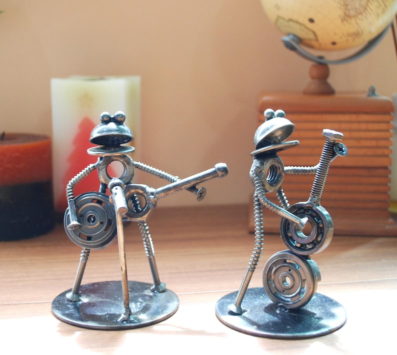 May include: Two metal frog sculptures playing musical instruments. One frog holds a microphone and a guitar, while the other plays a guitar. The sculptures are made of nuts, bolts, and washers, and are mounted on circular bases.