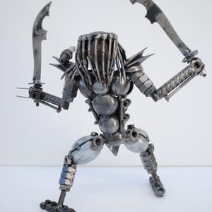 May include: A metal sculpture of a Predator from the movie franchise. The sculpture is made of various metal parts, including nuts, bolts, and screws. The Predator is holding two blades in its hands and is in a fighting stance.