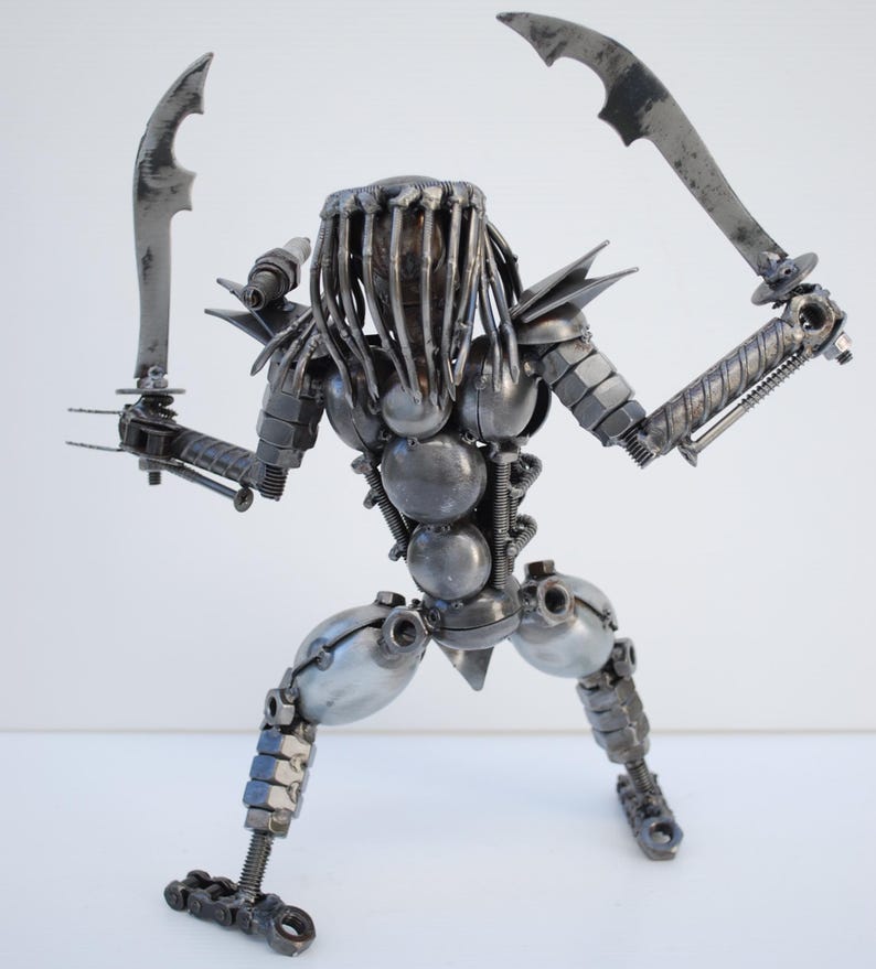May include: A metal sculpture of a Predator from the movie franchise. The sculpture is made of various metal parts, including nuts, bolts, and screws. The Predator is holding two blades in its hands and is in a fighting stance.