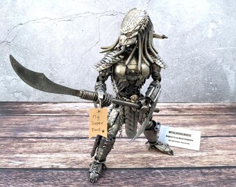 May include: A silver metal Predator sculpture with a large blade and a smaller knife. The sculpture is made from scrap metal and has detailed features. A tag reads "My Super Dad".