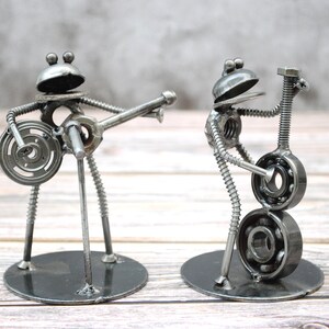 May include: Two metal frog sculptures playing musical instruments. One frog plays a guitar, the other a bass. The sculptures are made from metal nuts, bolts, and washers, and stand on round bases. The frogs have round heads and are painted silver.