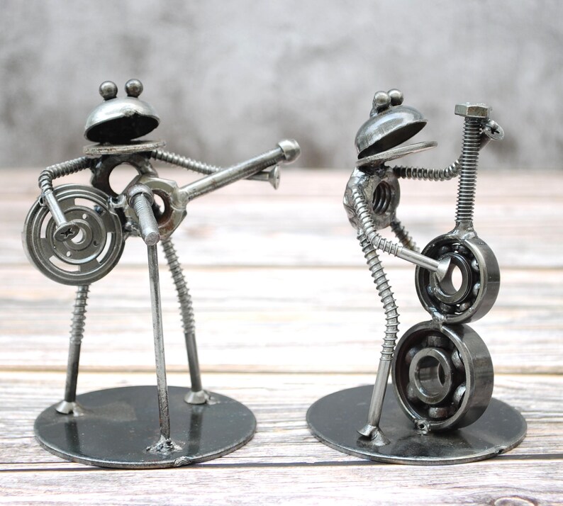 May include: Two metal frog sculptures playing musical instruments. One frog plays a guitar, the other a bass. The sculptures are made from metal nuts, bolts, and washers, and stand on round bases. The frogs have round heads and are painted silver.