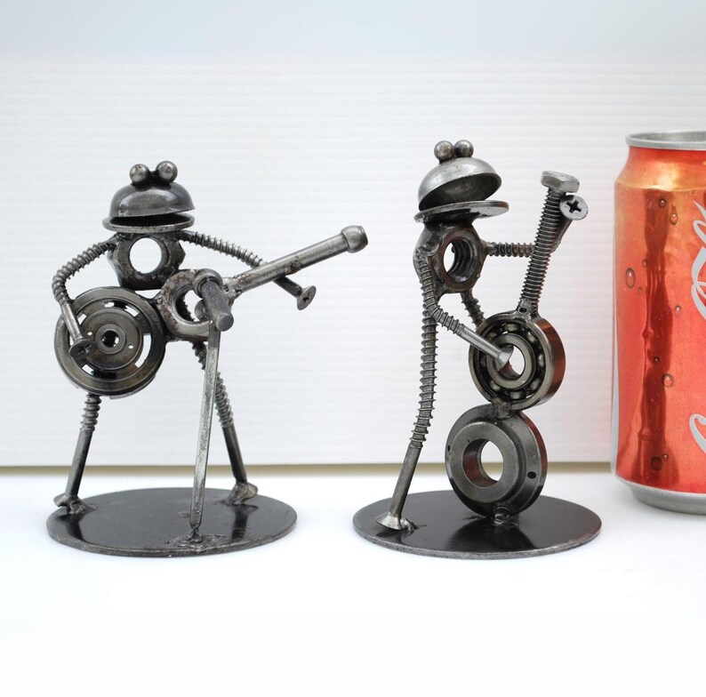 May include: Two metal frog sculptures playing musical instruments. One frog plays a guitar, the other a trumpet. The sculptures are made from nuts, bolts, and other metal hardware. A red soda can is next to the sculptures.