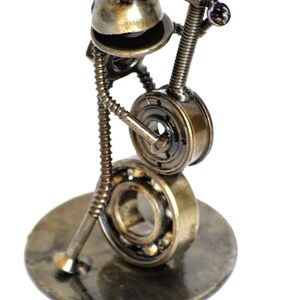 May include: A whimsical sculpture of a frog playing a banjo, crafted from repurposed metal hardware. The frog's head is a rounded piece, with a body made of a spring and screws. The banjo is made from a bearing and other metal parts, all mounted on a circular base.
