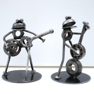May include: Two metal sculptures of musicians. One figure plays a guitar, the other a bass. The sculptures are made from nuts, bolts, and other metal hardware. Each figure stands on a round black base.
