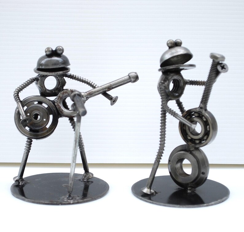 May include: Two metal sculptures of musicians. One figure plays a guitar, the other a bass. The sculptures are made from nuts, bolts, and other metal hardware. Each figure stands on a round black base.