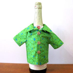 May include: A green short-sleeved shirt cover for a bottle, featuring a repeating blue and green floral pattern, a pointed collar, and three light brown wooden buttons down the center front. The shirt is fitted around the neck of a glass bottle.