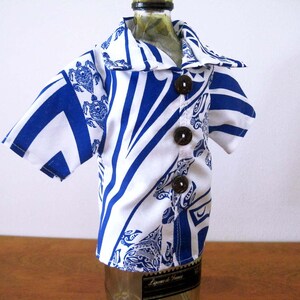 May include: A miniature short-sleeved shirt designed to fit over a bottle, featuring a white and blue geometric pattern with turtle motifs. The garment includes a pointed collar and three dark brown circular buttons down the front center.