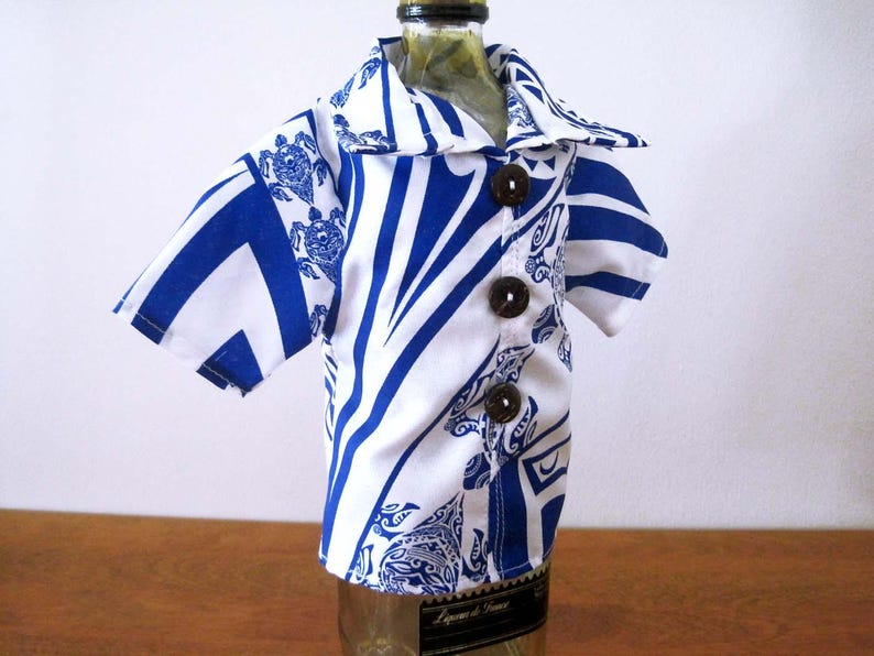 May include: A miniature short-sleeved shirt designed to fit over a bottle, featuring a white and blue geometric pattern with turtle motifs. The garment includes a pointed collar and three dark brown circular buttons down the front center.