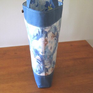 Puede incluir: Un bolso tote alto con la parte superior e inferior de mezclilla azul. El cuerpo principal de la bolsa presenta un estampado de una escena invernal con una combinaci&oacute;n de colores azul claro y blanco. La bolsa tiene dos asas y un cierre de bot&oacute;n de madera.