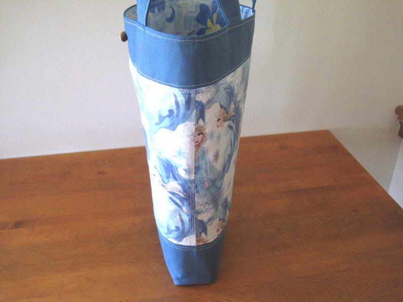 Puede incluir: Un bolso tote alto con la parte superior e inferior de mezclilla azul. El cuerpo principal de la bolsa presenta un estampado de una escena invernal con una combinaci&oacute;n de colores azul claro y blanco. La bolsa tiene dos asas y un cierre de bot&oacute;n de madera.