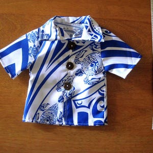 May include: A short-sleeved button-down shirt featuring a white and blue tribal pattern with sea turtle motifs and three dark brown buttons. A white measuring tape is positioned to the right, showing the shirt is approximately 8 inches long.