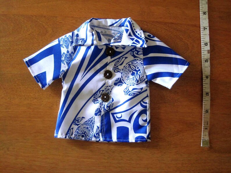 May include: A short-sleeved button-down shirt featuring a white and blue tribal pattern with sea turtle motifs and three dark brown buttons. A white measuring tape is positioned to the right, showing the shirt is approximately 8 inches long.