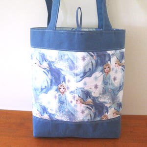 Puede incluir: Bolso tote azul con un dise&ntilde;o estampado que presenta a una princesa con un vestido azul, rodeada de copos de nieve y un fondo azul arremolinado. El bolso tiene asas azules y una base azul. El bolso est&aacute; hecho de tela.