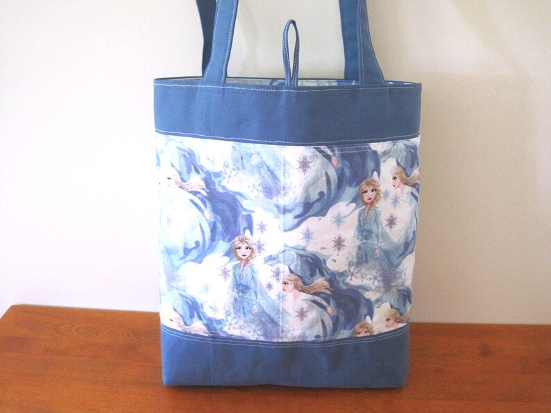 Puede incluir: Bolso tote azul con un dise&ntilde;o estampado que presenta a una princesa con un vestido azul, rodeada de copos de nieve y un fondo azul arremolinado. El bolso tiene asas azules y una base azul. El bolso est&aacute; hecho de tela.