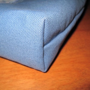 Puede incluir: Primer plano de una esquina de una bolsa de tela azul claro. La bolsa tiene un tejido texturizado y est&aacute; construida con costuras precisas. La esquina est&aacute; bien definida, mostrando la forma y la construcci&oacute;n de la bolsa. La bolsa es probablemente un bolso tote o similar.