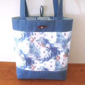 Puede incluir: Un bolso tote azul con un panel blanco con un patr&oacute;n repetido de una princesa con un vestido fluido, copos de nieve y un castillo. El bolso tiene asas y ribetes azules, y un cierre de palanca de madera.