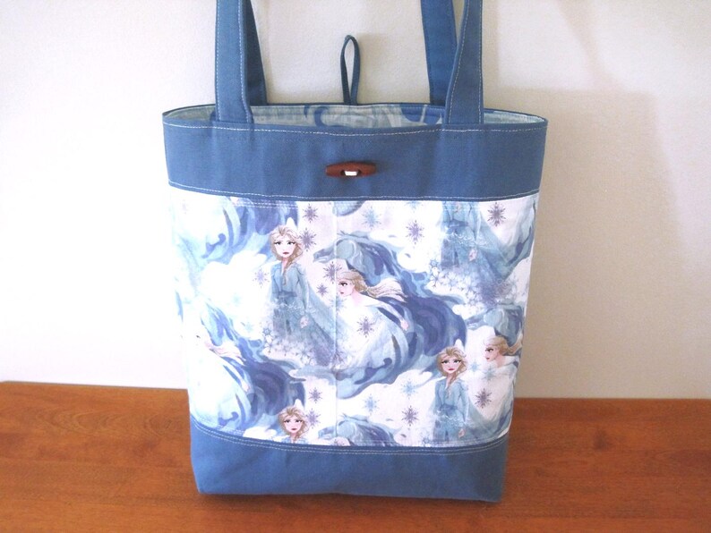 Puede incluir: Un bolso tote azul con un panel blanco con un patr&oacute;n repetido de una princesa con un vestido fluido, copos de nieve y un castillo. El bolso tiene asas y ribetes azules, y un cierre de palanca de madera.