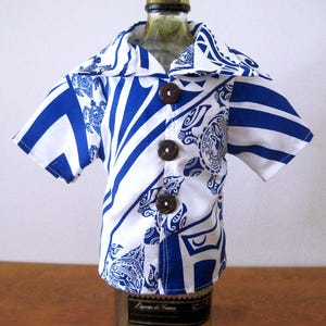 May include: A short-sleeved button-down shirt featuring a white and blue patterned design with sea turtle motifs. The garment has a wide collar and three dark brown circular buttons down the front, fitted over a glass bottle.
