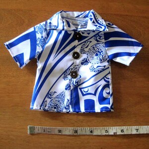 May include: A short-sleeved button-down shirt featuring a blue and white tribal pattern with sea turtle motifs. The shirt has three dark brown buttons and is positioned above a measuring tape showing a width of approximately 9 inches.