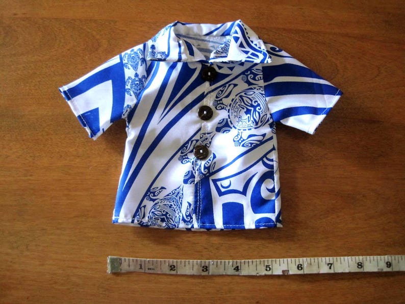 May include: A short-sleeved button-down shirt featuring a blue and white tribal pattern with sea turtle motifs. The shirt has three dark brown buttons and is positioned above a measuring tape showing a width of approximately 9 inches.