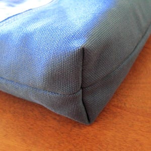 Puede incluir: Primer plano de una esquina de un bolso tote de lona azul. El bolso est&aacute; hecho de una tela resistente y texturizada con costuras visibles. El color es un azul apagado, con un toque de gris. El bolso parece estar vac&iacute;o.