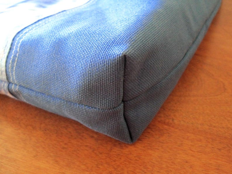 Puede incluir: Primer plano de una esquina de un bolso tote de lona azul. El bolso est&aacute; hecho de una tela resistente y texturizada con costuras visibles. El color es un azul apagado, con un toque de gris. El bolso parece estar vac&iacute;o.