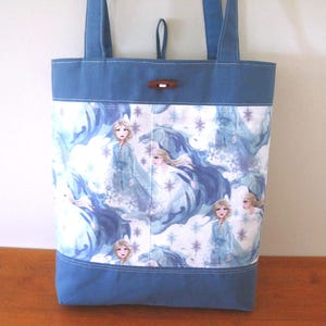 Puede incluir: Un bolso tote azul con un panel blanco con un patr&oacute;n repetido de un personaje con un vestido azul. El bolso tiene asas azules y un cierre de palanca de madera. El bolso est&aacute; hecho de tela.