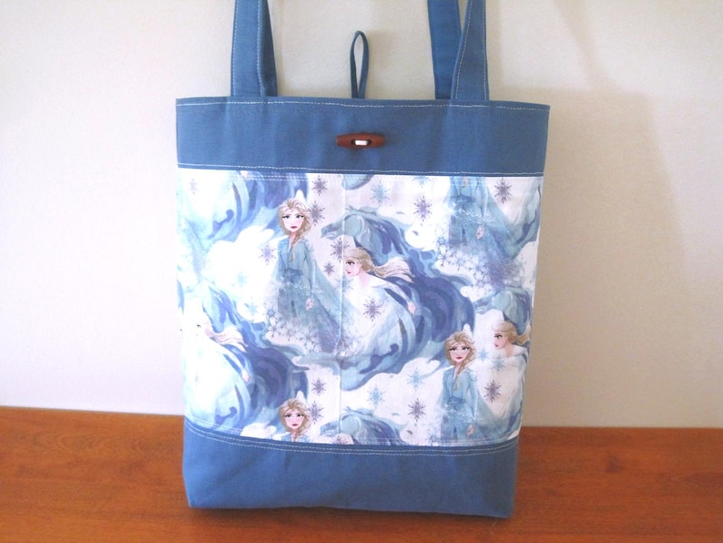 Puede incluir: Un bolso tote azul con un panel blanco con un patr&oacute;n repetido de un personaje con un vestido azul. El bolso tiene asas azules y un cierre de palanca de madera. El bolso est&aacute; hecho de tela.