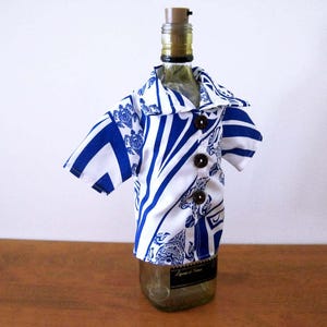 May include: A glass bottle wearing a short-sleeved shirt cover featuring a white and blue geometric pattern with sea turtle motifs. The shirt has a pointed collar and three dark brown circular buttons down the front center, secured with a cork stopper.