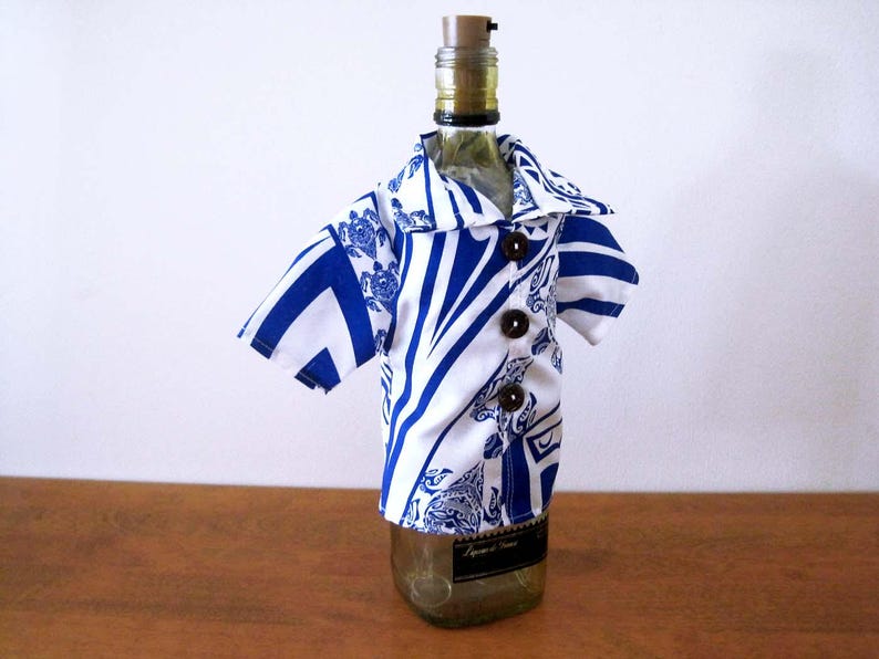 May include: A glass bottle wearing a short-sleeved shirt cover featuring a white and blue geometric pattern with sea turtle motifs. The shirt has a pointed collar and three dark brown circular buttons down the front center, secured with a cork stopper.
