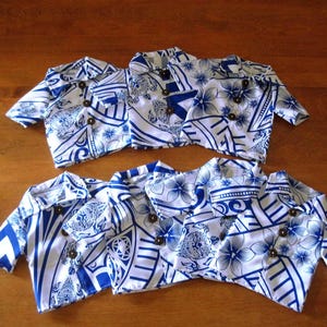 May include: Six short-sleeved button-down shirts featuring a white and blue floral and geometric pattern. Each shirt has a pointed collar and three dark brown circular buttons down the front. The shirts are arranged in two rows of three on a surface.