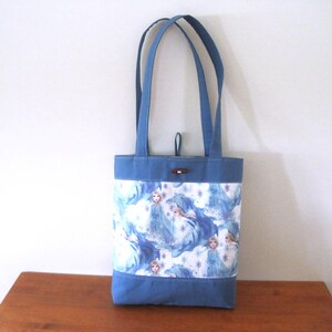 Puede incluir: Bolso tote azul con un estampado blanco y azul con un dise&ntilde;o de princesa. El bolso tiene asas azules y una base azul. Un bot&oacute;n de madera asegura la parte superior. Aproximadamente 30 cm de alto.