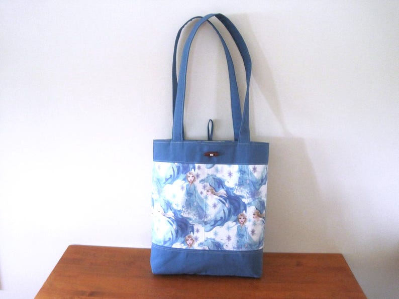 Puede incluir: Bolso tote azul con un estampado blanco y azul con un dise&ntilde;o de princesa. El bolso tiene asas azules y una base azul. Un bot&oacute;n de madera asegura la parte superior. Aproximadamente 30 cm de alto.