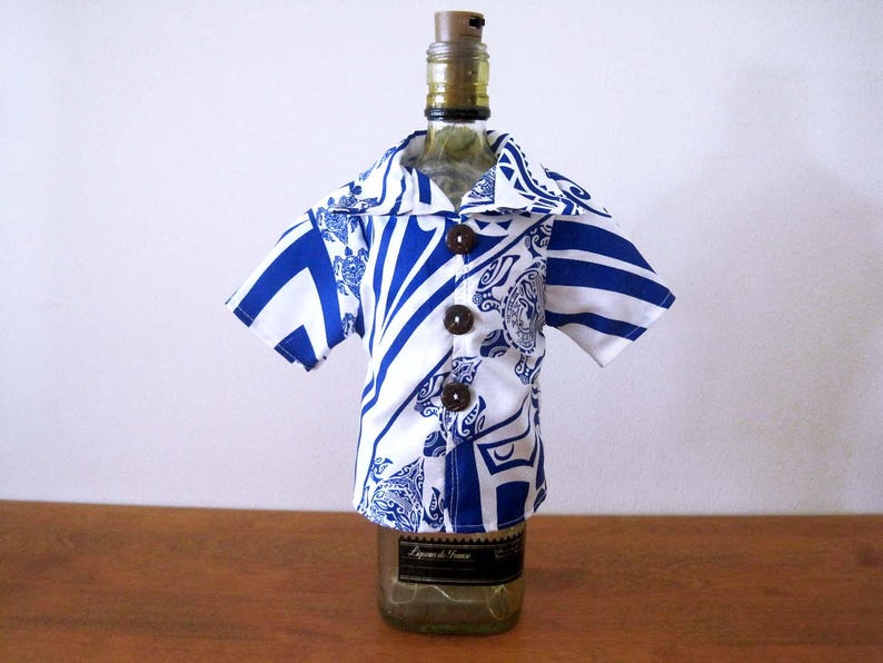 May include: A short-sleeved button-down shirt cover for a bottle, featuring a white and blue geometric and floral pattern. The garment has a wide collar and three dark brown circular buttons down the front, fitted over a glass bottle.