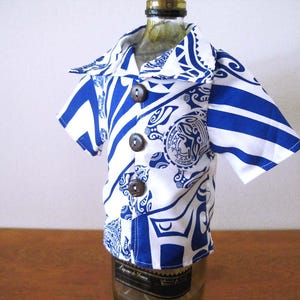 May include: A short-sleeved button-down shirt in white with a bold blue graphic pattern featuring tribal-inspired motifs and circular emblems. The garment has a wide collar and three dark brown circular buttons down the front center placket.