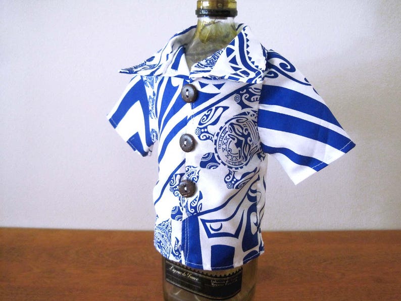 May include: A short-sleeved button-down shirt in white with a bold blue graphic pattern featuring tribal-inspired motifs and circular emblems. The garment has a wide collar and three dark brown circular buttons down the front center placket.