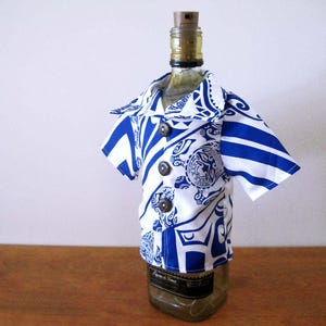 May include: A glass bottle wearing a short-sleeved shirt with a blue and white tribal-inspired pattern. The shirt features a collared neckline, three dark brown buttons down the front, and a graphic print that includes a circular emblem design.