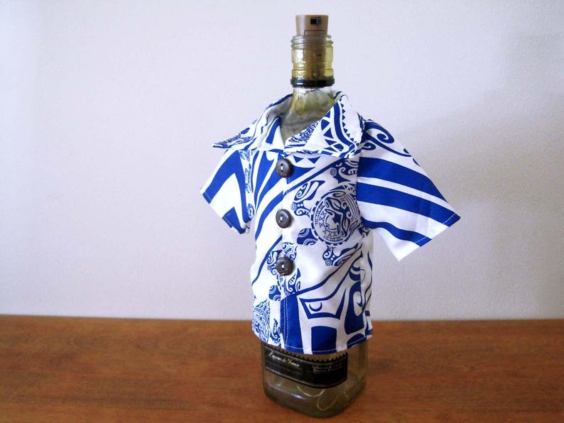 May include: A glass bottle wearing a short-sleeved shirt with a blue and white tribal-inspired pattern. The shirt features a collared neckline, three dark brown buttons down the front, and a graphic print that includes a circular emblem design.