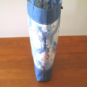 Puede incluir: Un bolso tote alto con la parte superior e inferior azul claro, y un centro estampado en blanco y azul. El estampado parece representar personajes de la pel&iacute;cula Frozen. El bolso est&aacute; sobre una superficie de madera.